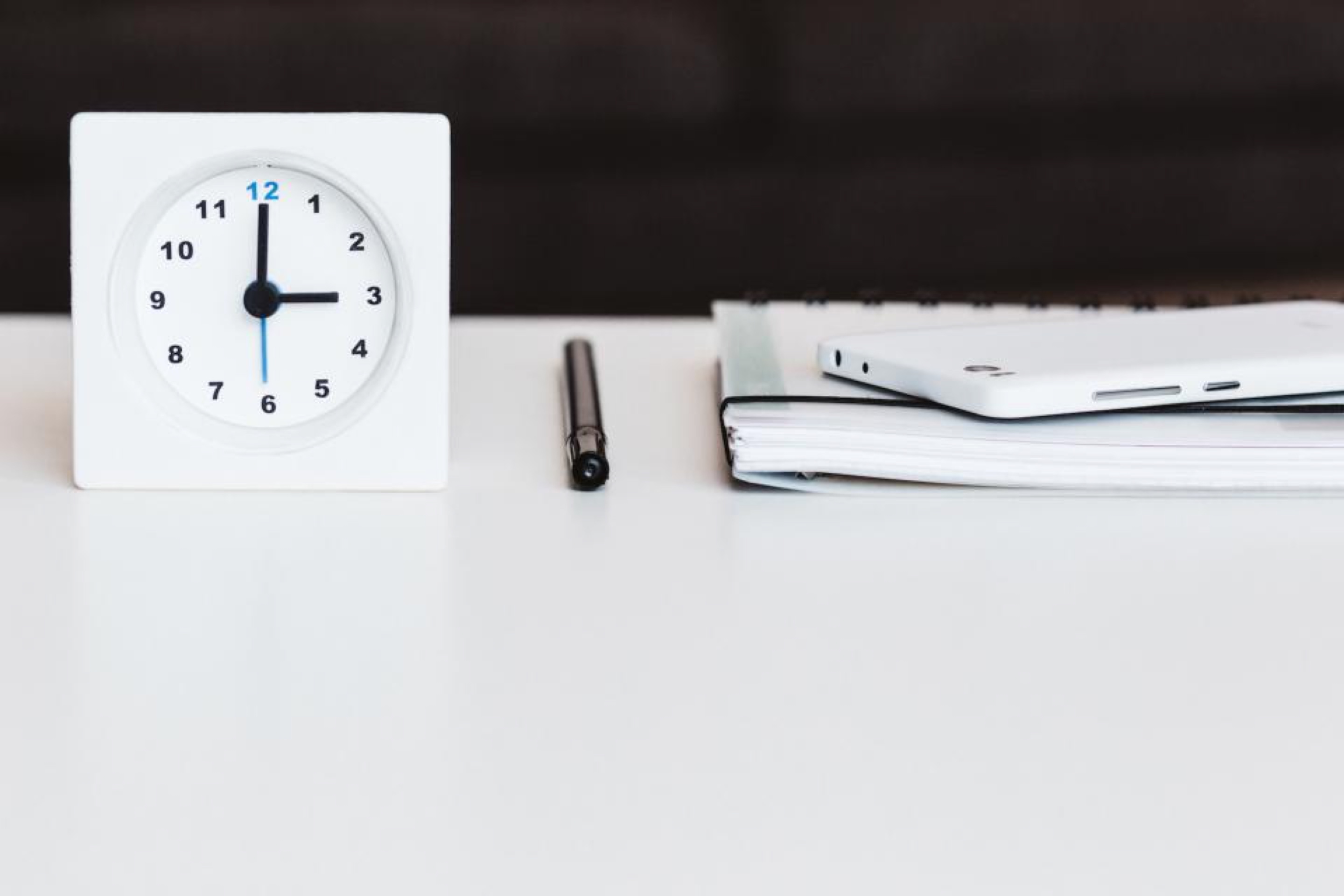 Petite horloge blanche posée sur un bureau blanc, à côté d'un stylo et d'un carnet.
